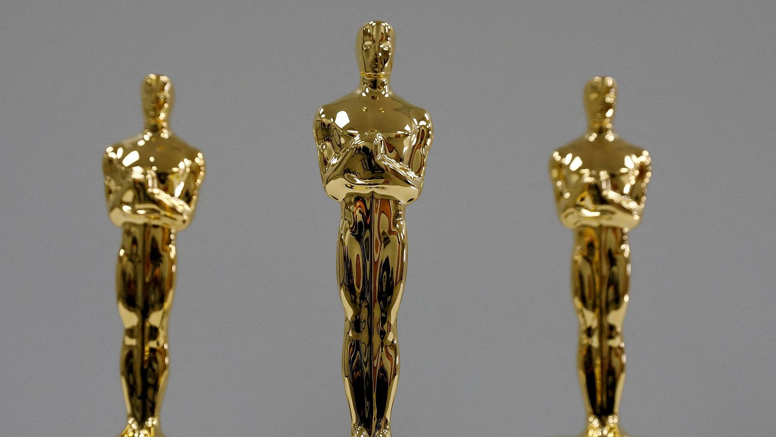 Tres estatuillas doradas de los Oscar, una central y dos laterales desenfocadas, sobre fondo gris.  Su brillo y forma son los protagonistas de la imagen.