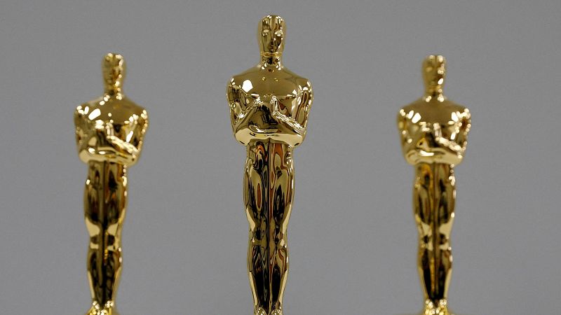 Tres estatuillas doradas de los Oscar, una central y dos laterales desenfocadas, sobre fondo gris.  Su brillo y forma son los protagonistas de la imagen.