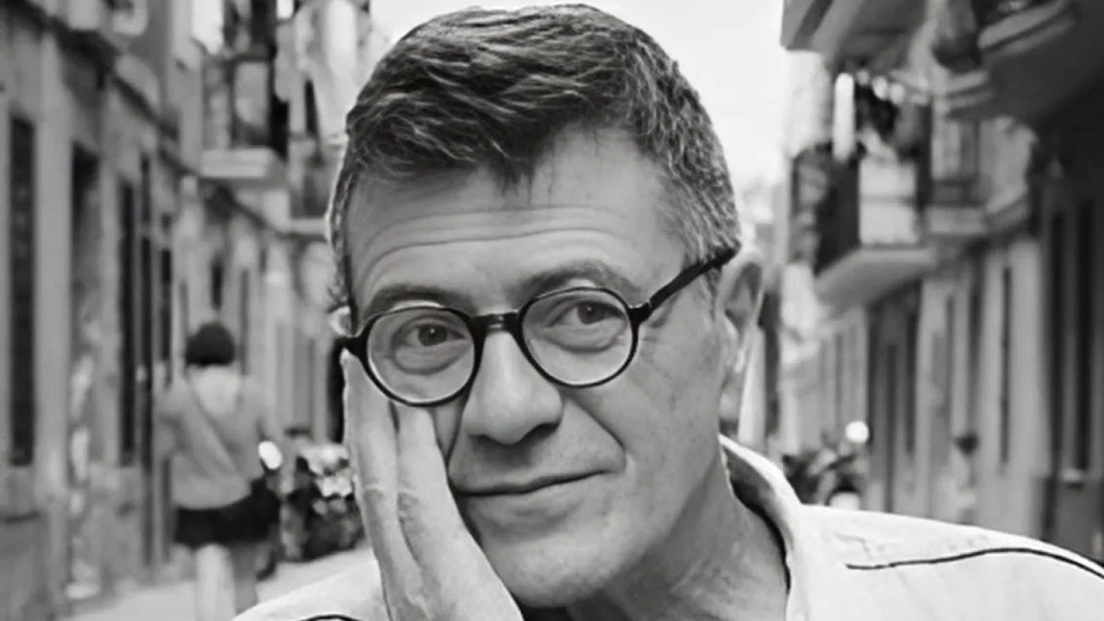 Retrato en blanco y negro de un hombre de mediana edad con gafas, expresión serena y mano izquierda en la mejilla; fondo urbano borroso.