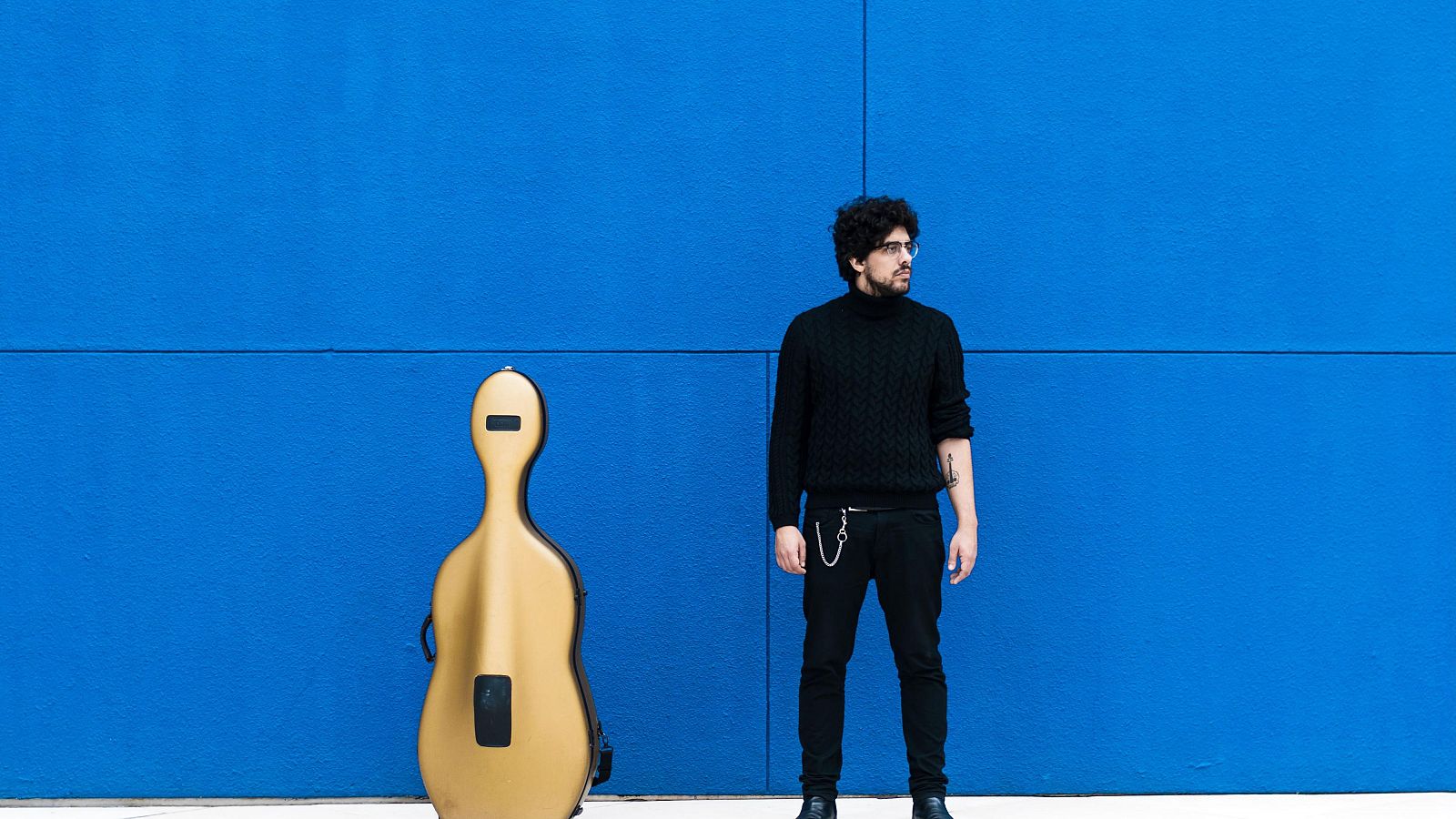 Hombre con gafas y tatuaje, vestido de negro, junto a un violonchelo en su estuche dorado, apoyado en una pared azul brillante.