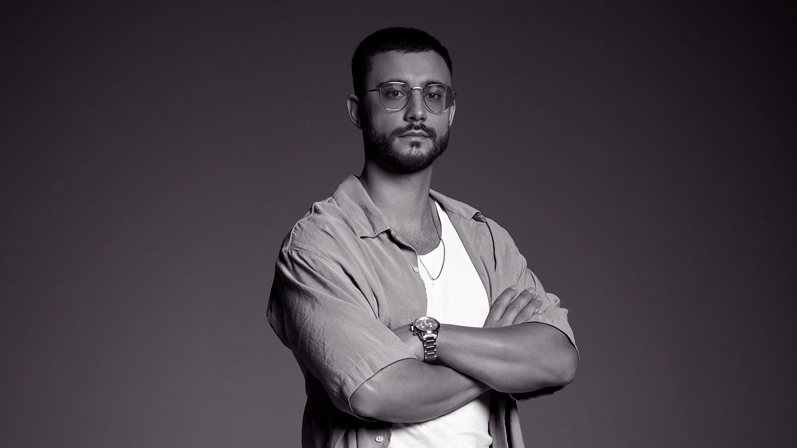 Retrato en blanco y negro de un hombre con gafas, camisa de manga corta, barba y expresión seria, mirando a cámara. Lleva reloj y cadena.