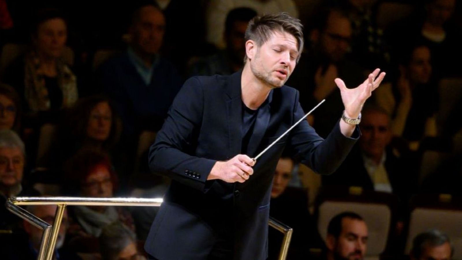 Director de orquesta concentrado dirigiendo una orquesta con batuta en mano; viste americana y camiseta negras. Público desenfocado al fondo.