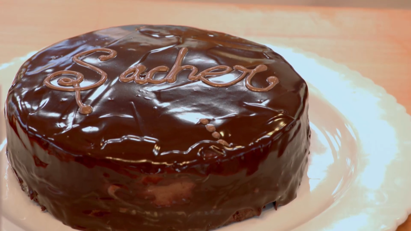 Tarta Sacher con glaseado de chocolate brillante, la palabra "Sacher" escrita en chocolate sobre el glaseado, presentada en un plato blanco.