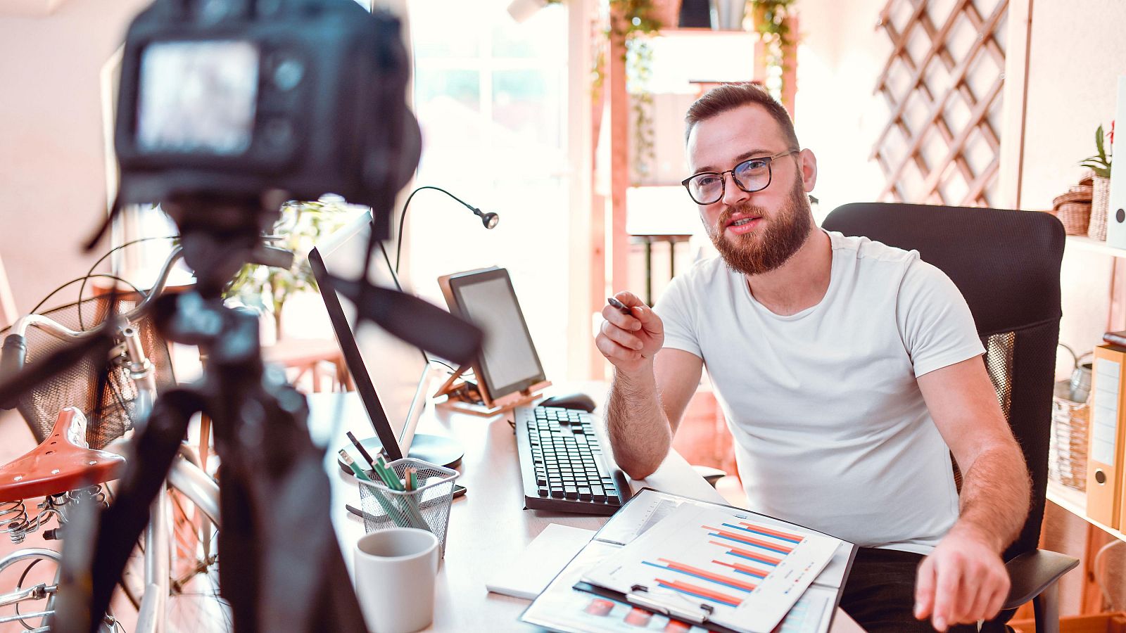 Hombre con barba y gafas graba vídeo explicativo señalando gráficos en su escritorio con dos monitores.  Utiliza bolígrafo y cámara en trípode.