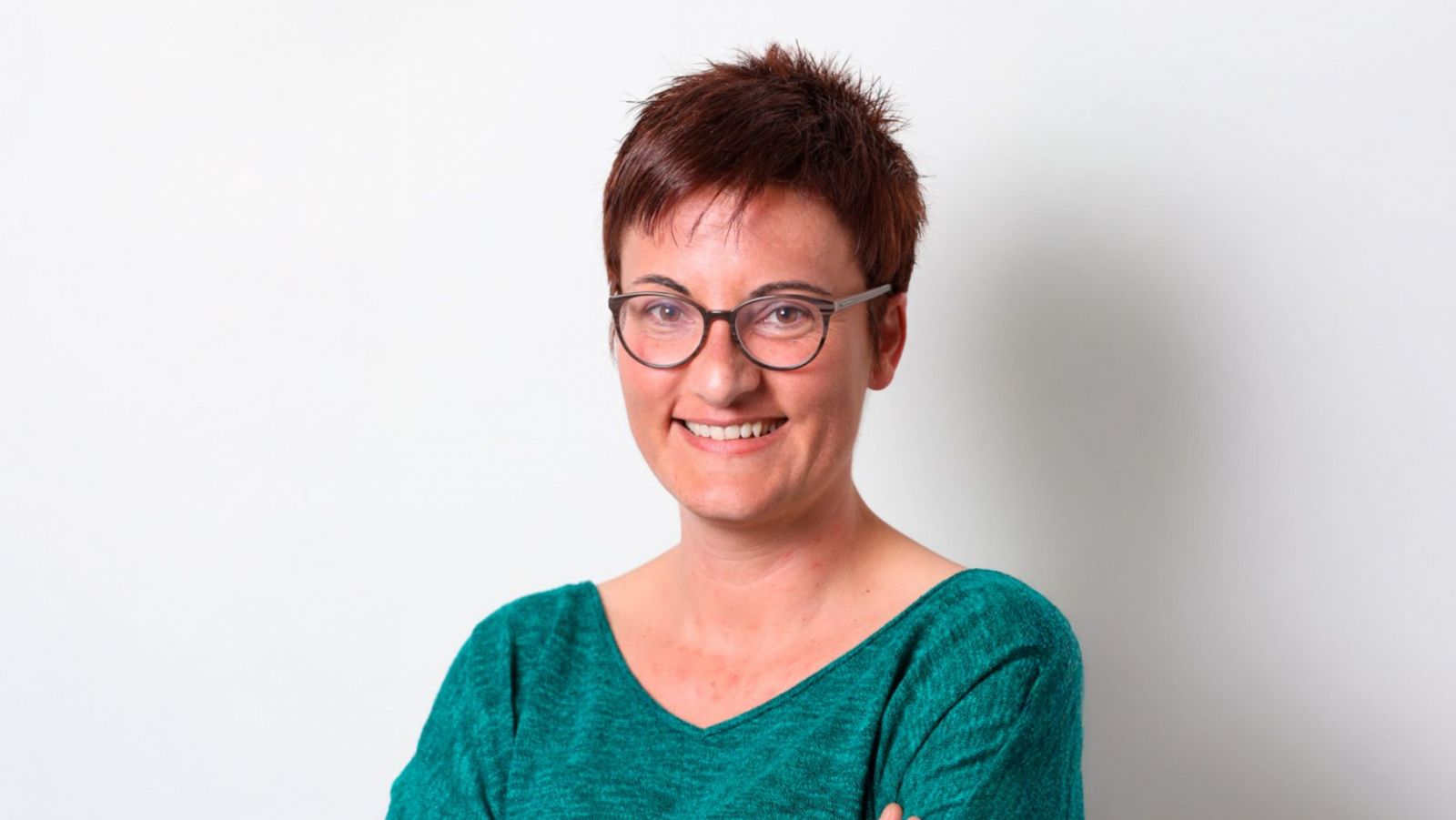 Retrato de una mujer con pelo corto rojizo, gafas y blusa verde oscuro, sonriendo a cámara sobre fondo blanco.