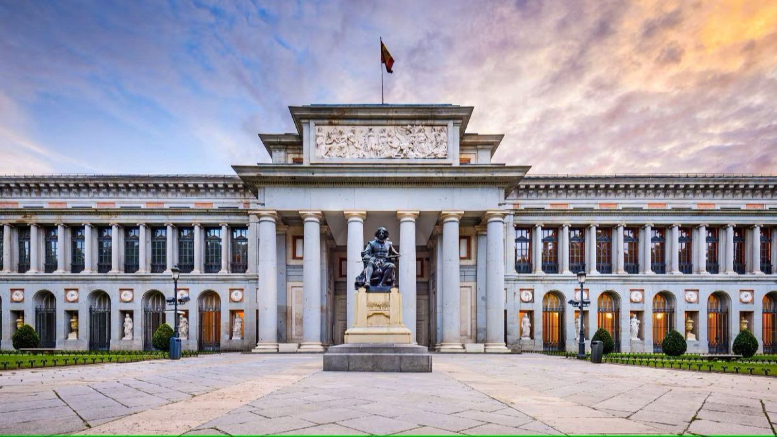 Fachada del Museo del Prado al amanecer o atardecer, con columnata, estatua y bandera española ondeando. Iluminación suave y cálida.