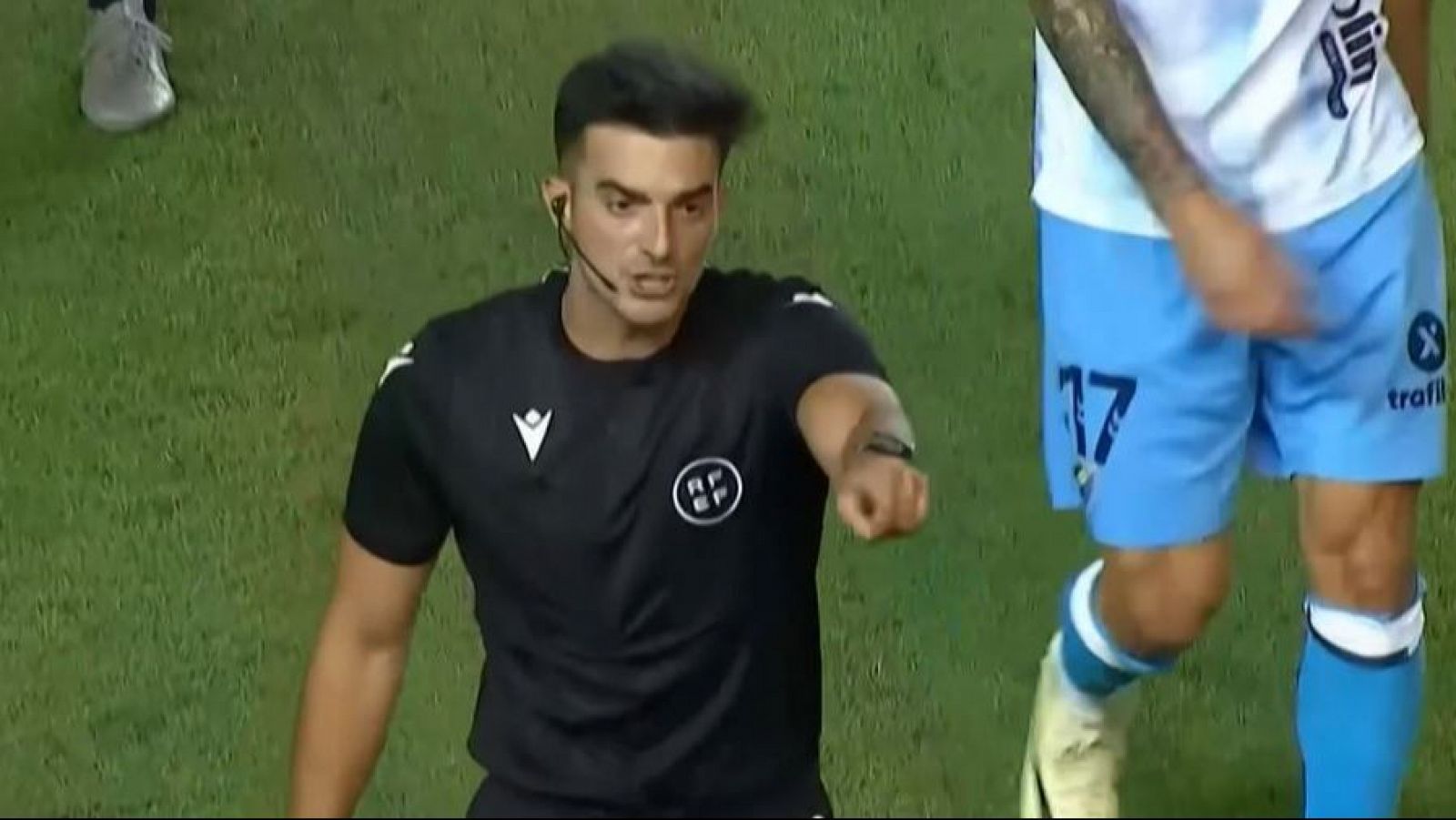 Árbitro con camiseta negra y logo RFEF señalando; a su derecha, jugador con pantalón corto azul y número 17.  Posiblemente relacionado con una disputa deportiva.