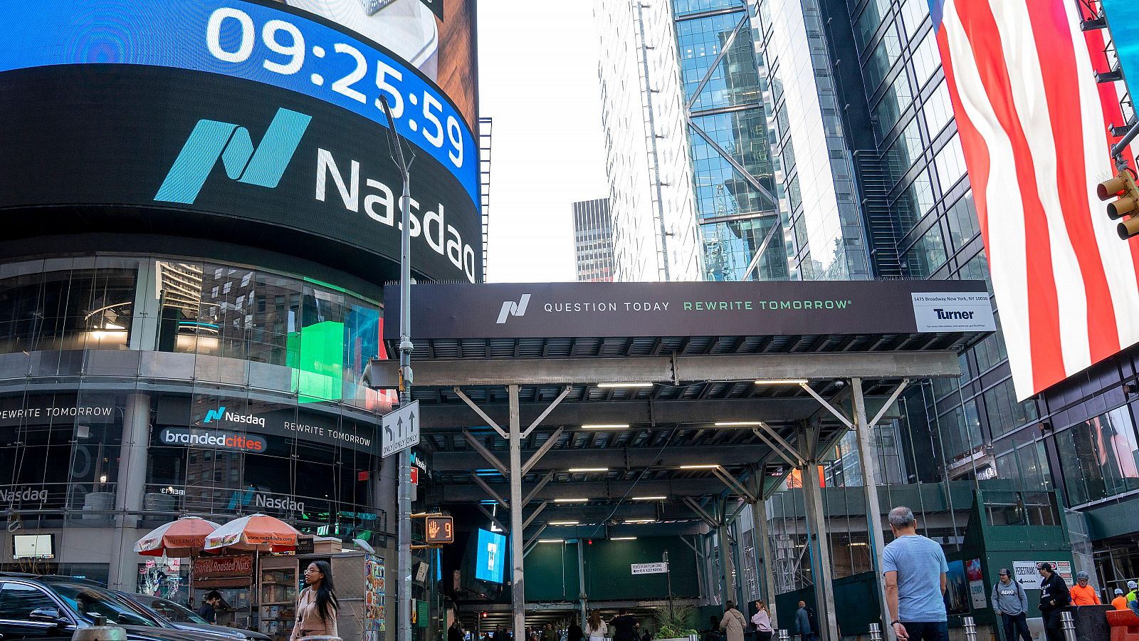 Pantalla Nasdaq en Times Square mostrando la hora 09:25:59. Andamio con el logo de Turner y el eslogan "QUESTION TODAY REWRITE TOMORROW".  Actividad urbana matutina.