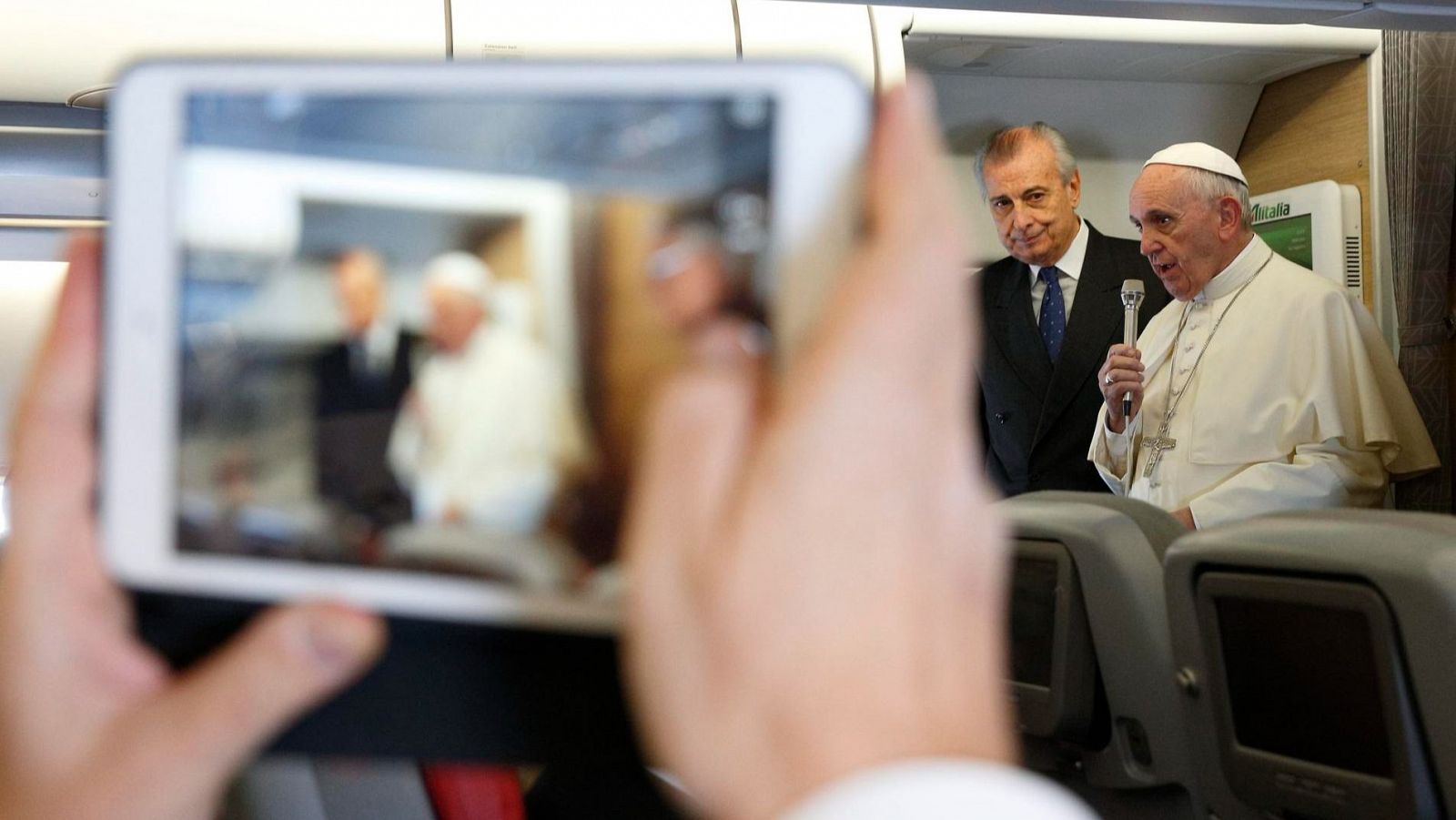 Enfocan al papa Francisco con una tableta mientras habla por un micrófono entre los asientos de un avión.