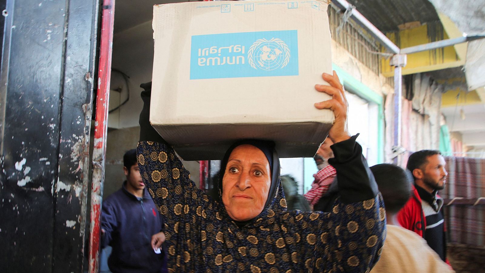 Mujer con caja UNRWA en la cabeza, expresión seria. Fondo con personas y estructuras metálicas, posible mercado o centro de ayuda.