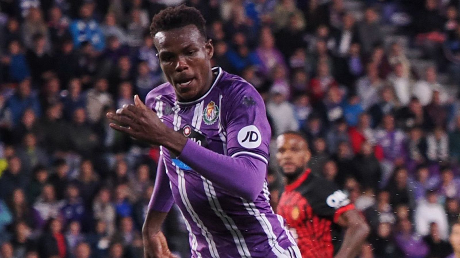 Jugador de fútbol con camiseta morada y blanca, logo JD en la manga, corriendo en el campo con expresión intensa. Fondo borroso con grada y otro jugador.