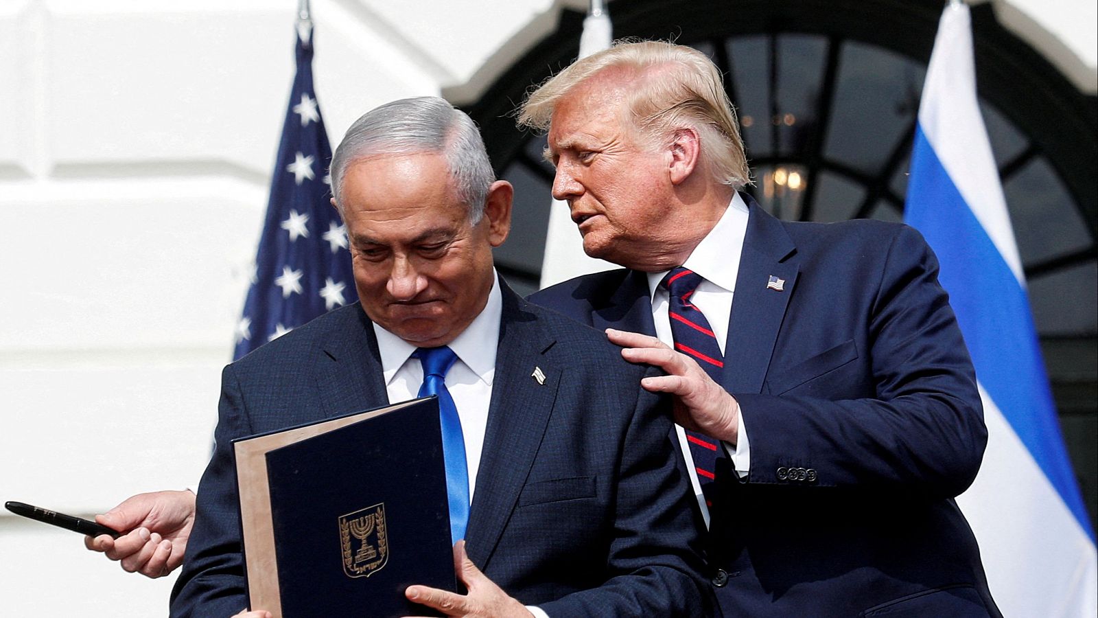 Reunión entre Donald Trump y Benjamin Netanyahu.  Netanyahu porta un documento con el escudo de Israel; Trump le tiene la mano en el hombro.  Banderas de EEUU e Israel al fondo.