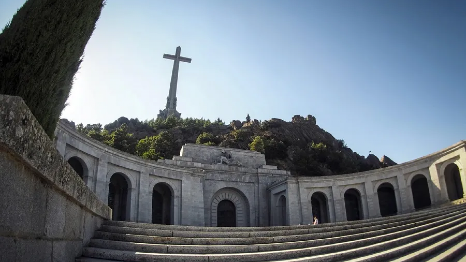 Fachada de basílica con cruz de piedra, escalinata y arcos en la entrada principal, coronada por escultura. Imagen de archivo.