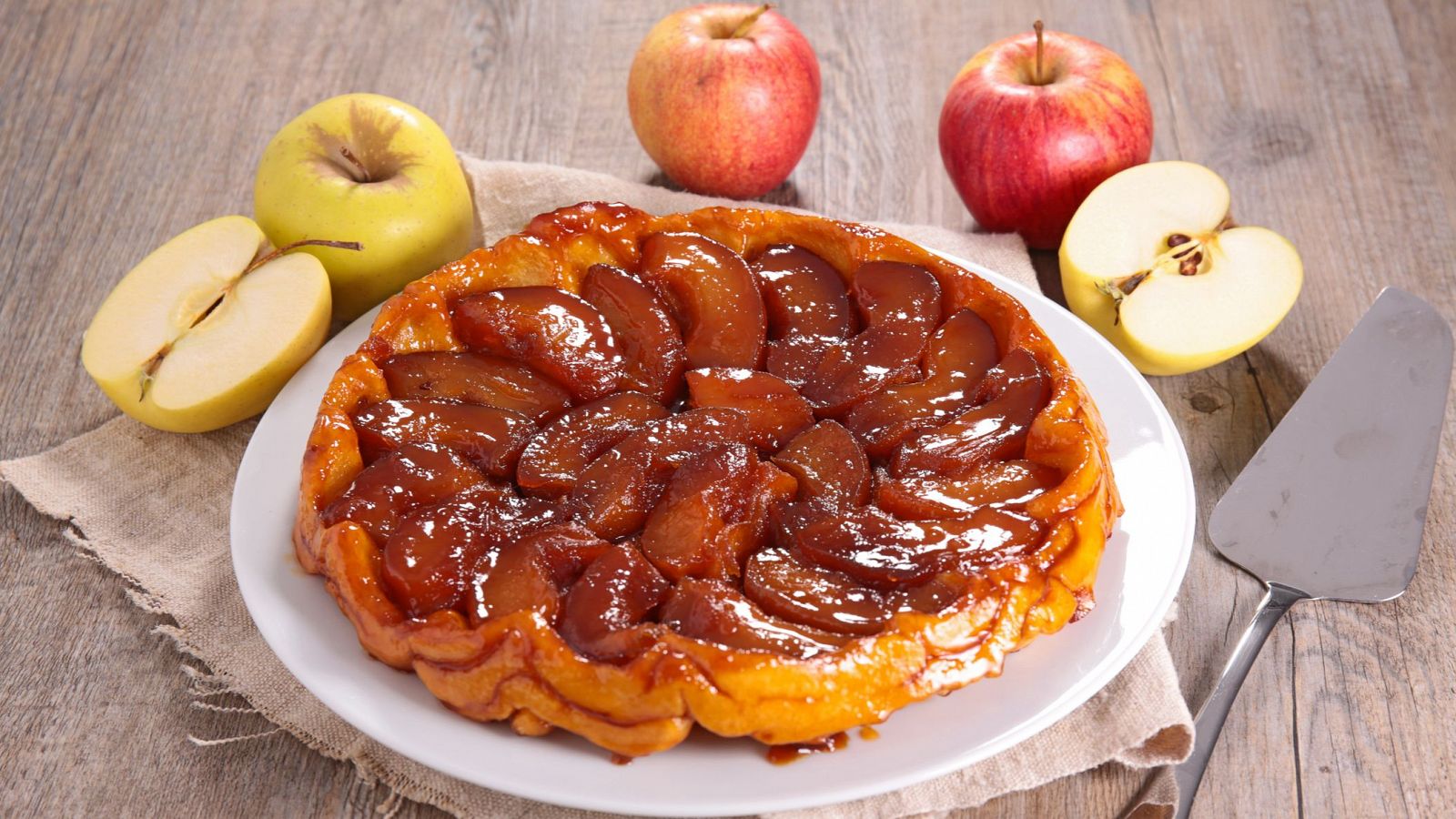 Tarta tatin con manzanas caramelizadas, servida en plato blanco junto a manzanas frescas y espátula.  Aspecto apetitoso y hogareño.