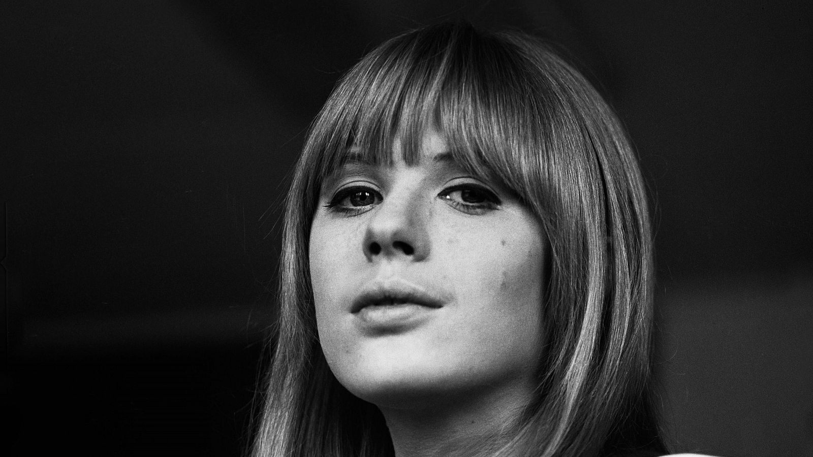 Retrato en blanco y negro de una joven con cabello liso hasta los hombros y flequillo, mirada hacia arriba y a la derecha. Fondo oscuro y desenfocado.