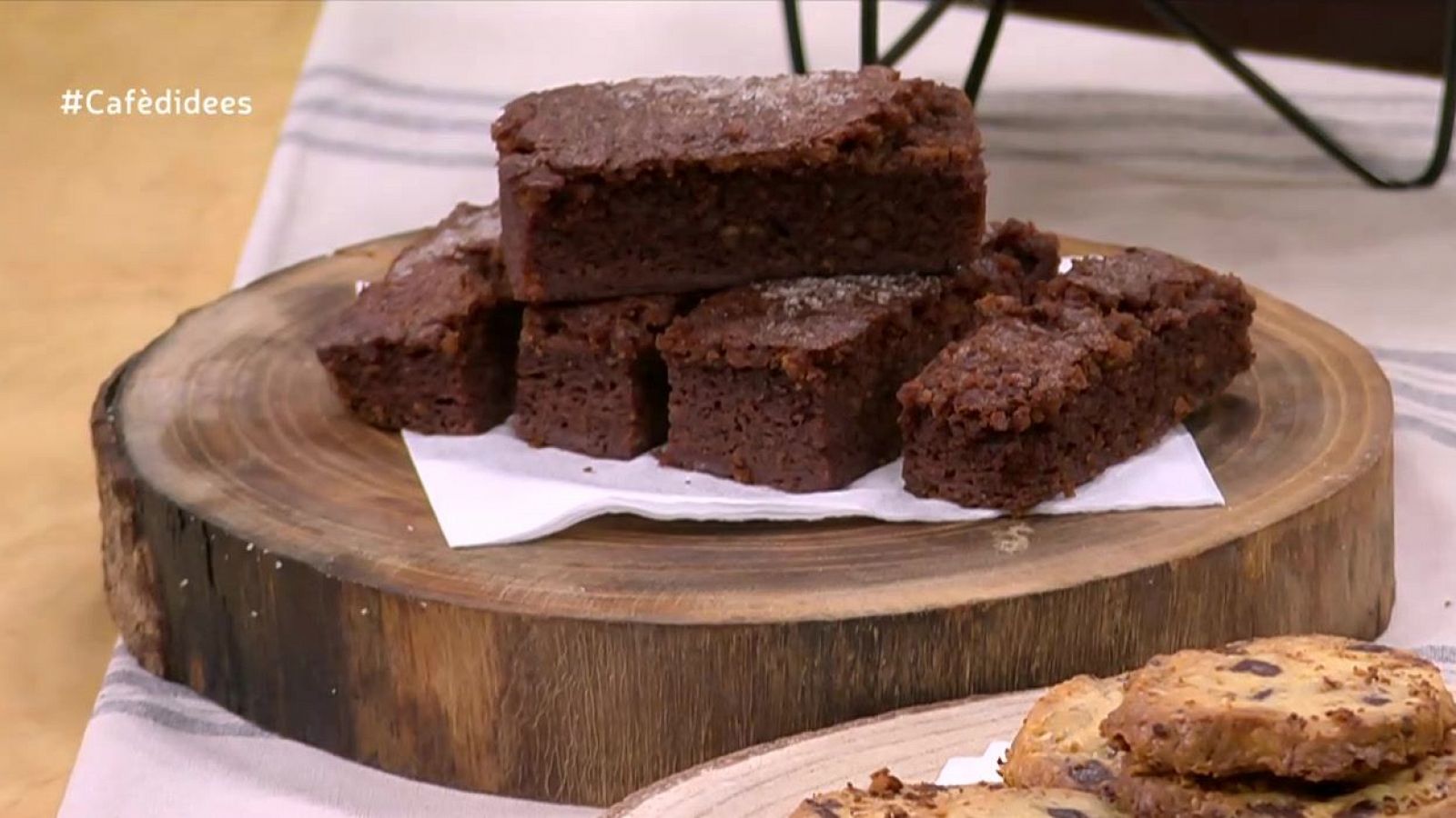 Brownies con azúcar, sobre tronco de madera y papel blanco, junto a galletas con pepitas de chocolate.  Incluye los textos '#Cafèdideees' y 'Pastís per recuperar galetes'.