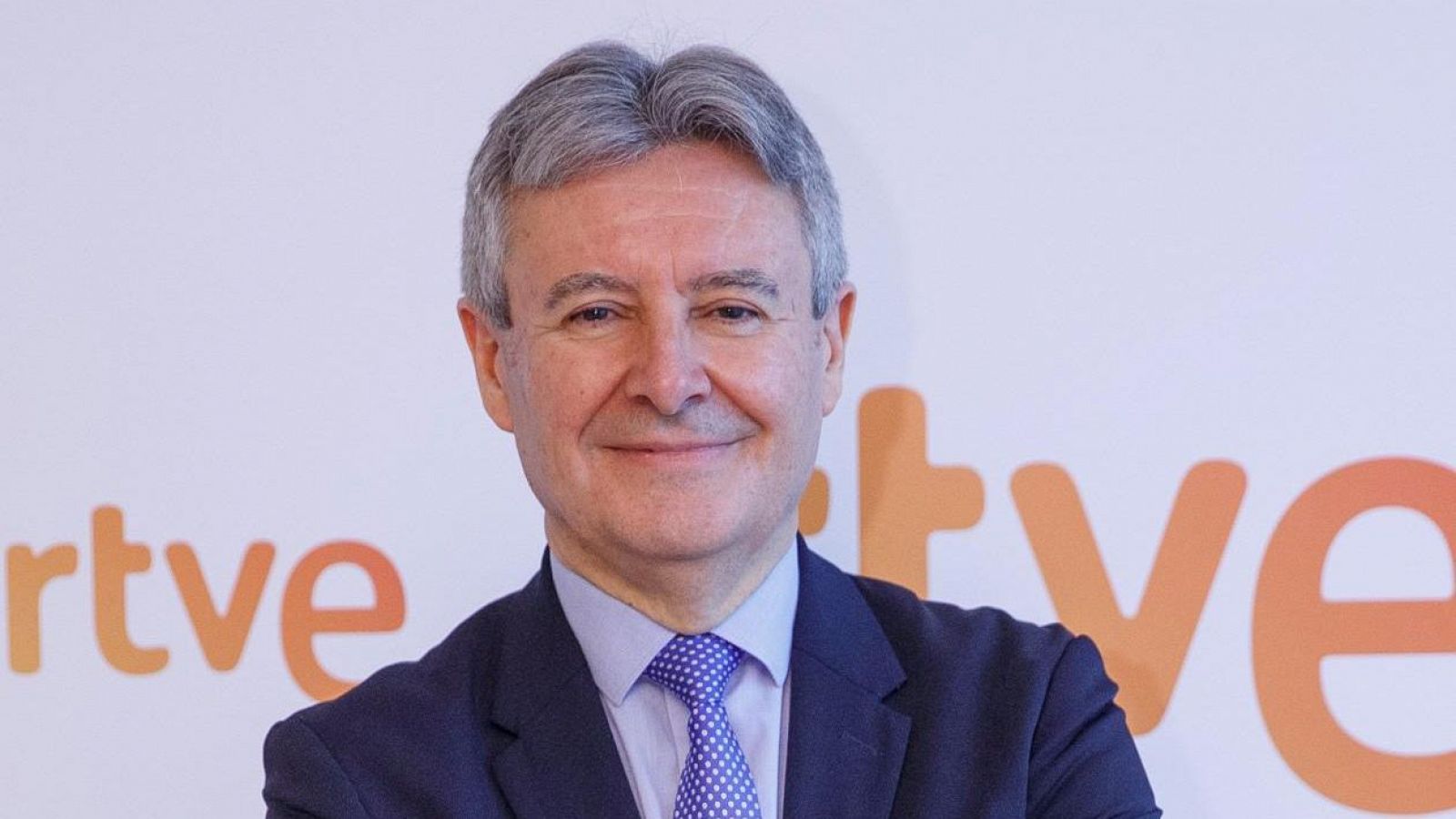 Retrato profesional de un hombre de mediana edad con camisa azul, corbata morada y chaqueta azul marino, sobre fondo blanco con logo RTVE.