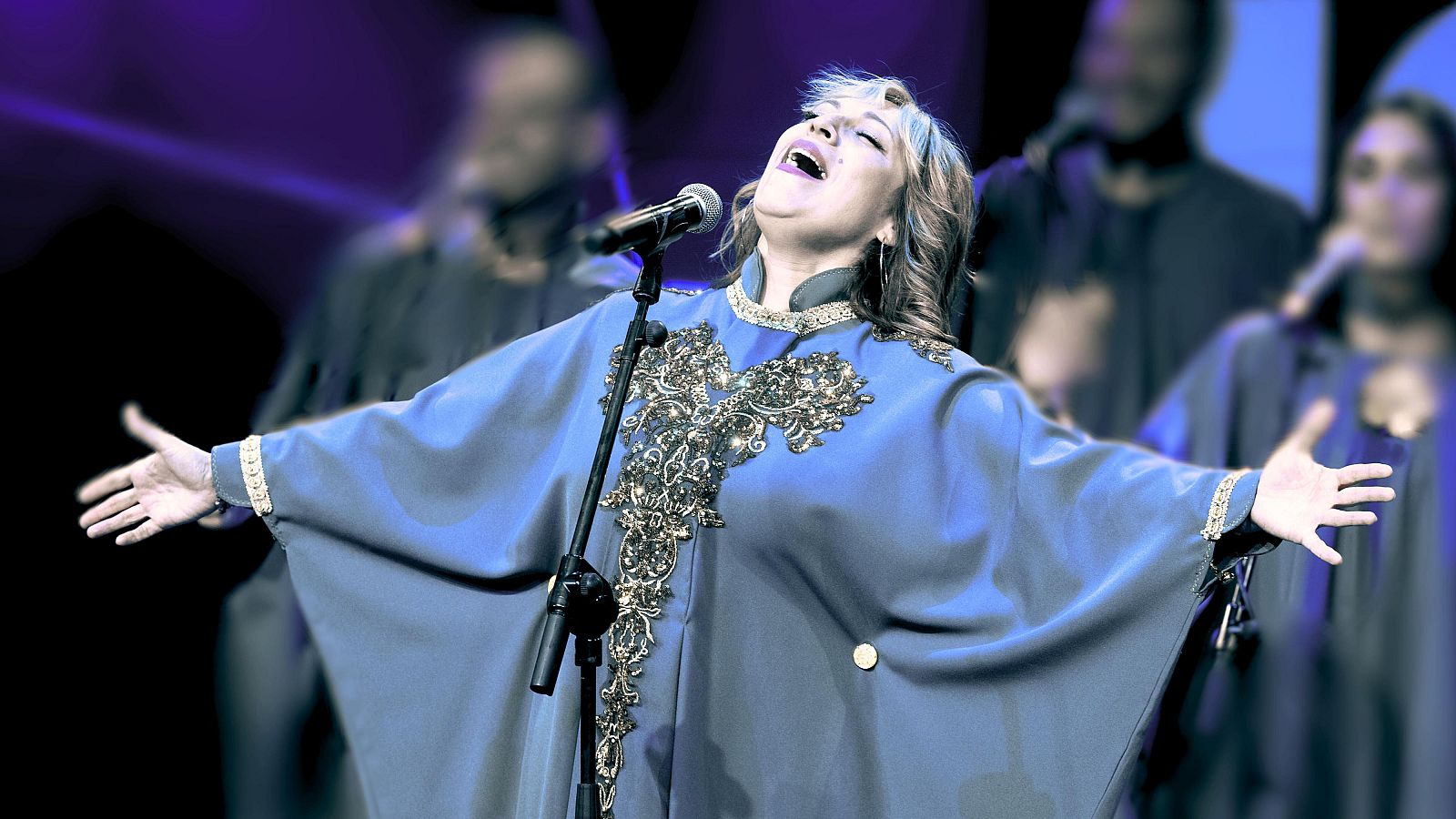 Cantante con túnica azul y bordados dorados en escena, micrófono delante, coro borroso al fondo. Iluminación destacando a la solista.