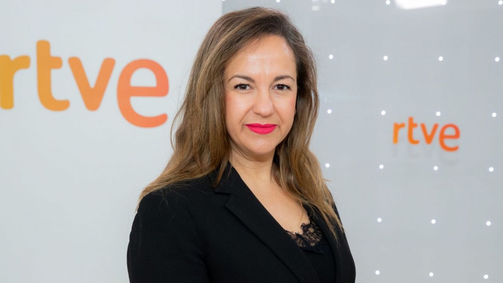 Retrato de mujer de mediana edad con cabello castaño claro, chaqueta negra y blusa oscura con encaje. Fondo blanco con logo RTVE y luces. Expresión seria.