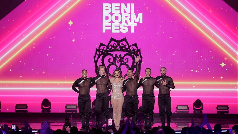 Cantante con traje brillante actuando en escenario rosa neón con cinco bailarines. Público entusiasmado en el Benidorm Fest.