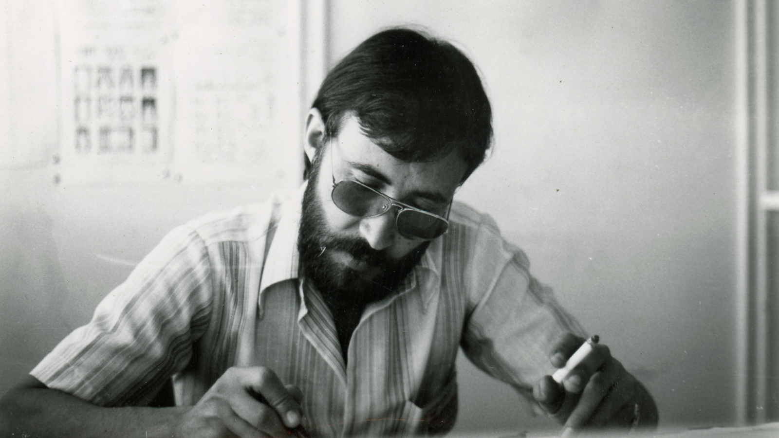 Hombre de mediana edad, con barba y gafas de sol, sentado a una mesa escribiendo o dibujando.  Imagen en blanco y negro con iluminación difusa.