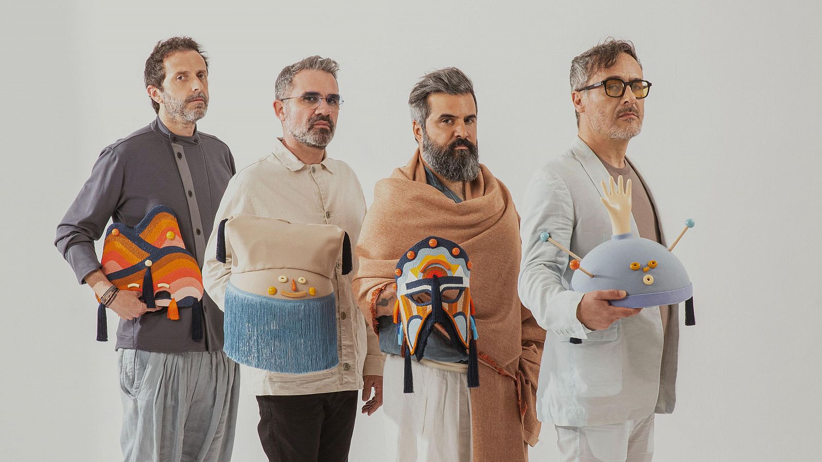 Cuatro músicos posan con objetos textiles coloridos y artesanales: alas de mariposa, pieza rectangular con cara sonriente, máscara facial y cúpula con antenas.