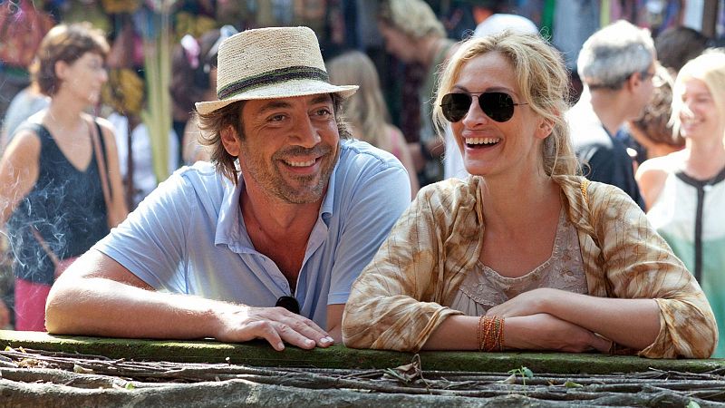 Escena de "Come, reza, ama" con Javier Bardem y Julia Roberts sonrientes, apoyados en una baranda en un ambiente cálido y público.