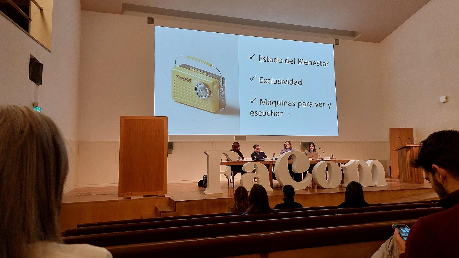 Sesión de clausura de congreso: ponentes en mesa, pantalla con radio antigua y conceptos relacionados con comunicación y bienestar.  Letras grandes "FaCom" en el escenario.