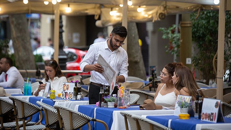 Terraza de restaurante con camarero mostrando la carta a clientes; mesas con bebidas y comida.  Contexto: noticia sobre tributación del salario mínimo.