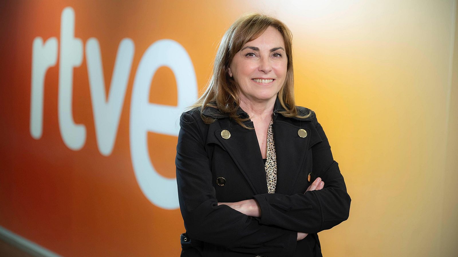 Retrato corporativo de una mujer de mediana edad con chaqueta negra y blusa estampada, frente a un fondo naranja con logo RTVE.