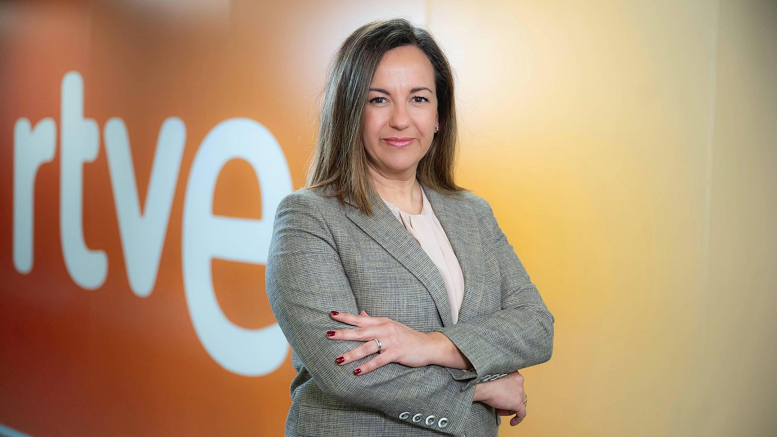 Directiva de RTVE con americana gris clara y blusa beige, frente al logo de la corporación. Expresión seria y amable.