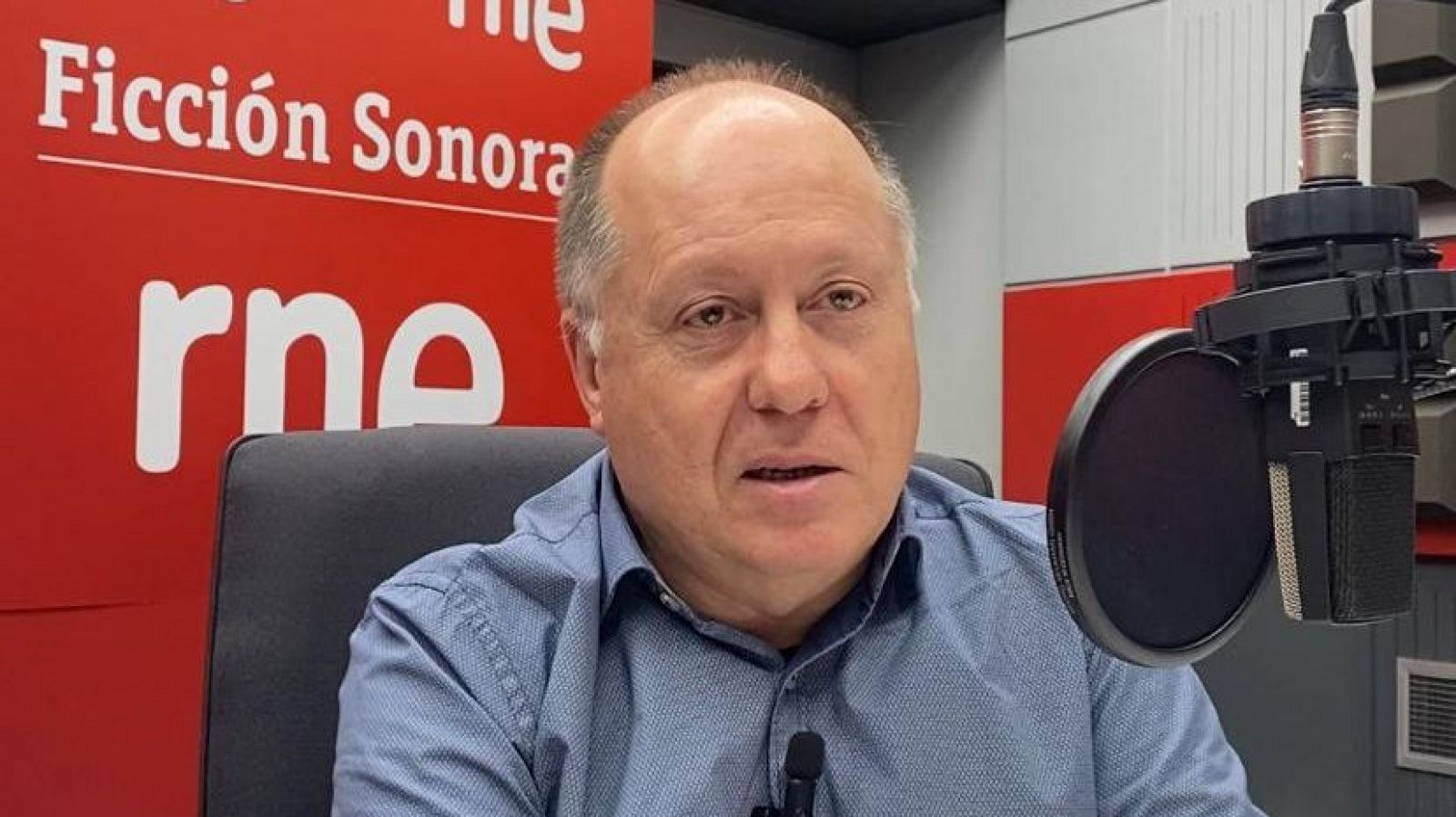 Imagen de Benigno Moreno en un estudio de grabación de RNE.  Viste camisa azul y se observa un micrófono y parte del logo de RNE y un cartel con "Ficción Sonora".