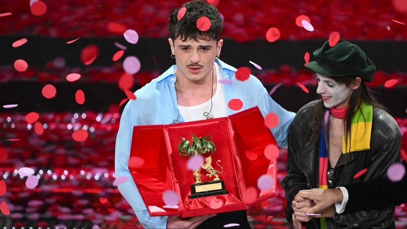 Olly, con camisa azul, celebra su victoria en Sanremo 2025 sosteniendo un trofeo en una caja roja.  Confetti rosa y rojo caen sobre él y otras personas.
