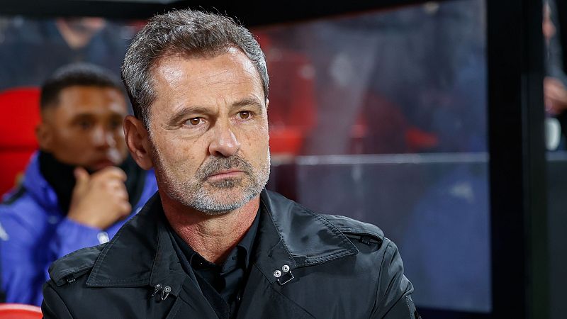 Imagen de Diego Cocca, ex entrenador, con chaqueta negra y expresión seria. Fondo desenfocado con asientos rojos y una persona con chaqueta azul.