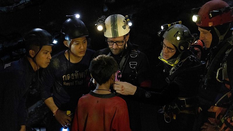 Grupo de personas con cascos y luces frontales atienden a un niño en una cueva oscura. Escena nocturna, posible rescate.