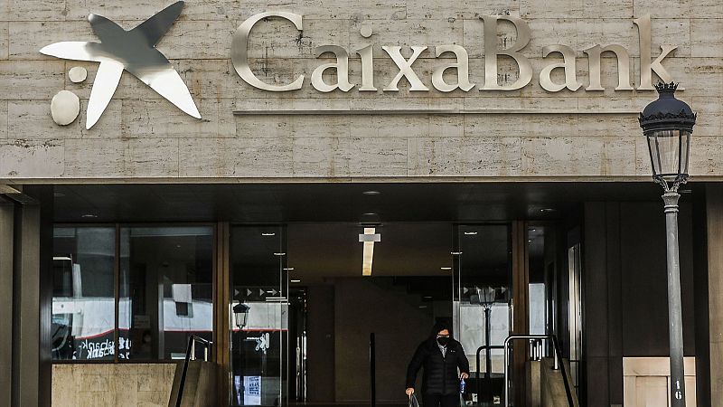 Entrada de una sucursal de CaixaBank con su logotipo visible. Una persona con mascarilla sale del edificio, junto a una farola clásica.