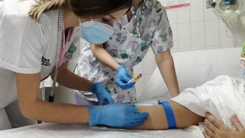 Extracción de sangre a paciente hospitalizado, asistido por dos enfermeras con mascarillas y uniformes.  Se observa una vía intravenosa.