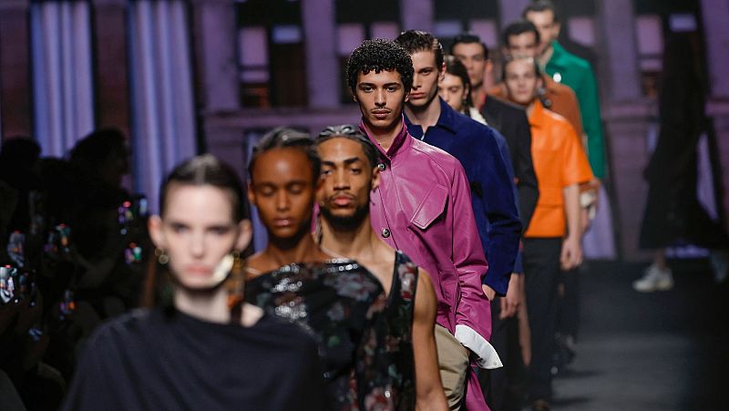 Modelos con atuendos diversos desfilan en una pasarela. Prendas en colores como fucsia, azul marino, naranja y verde destacan. Público observa el evento.