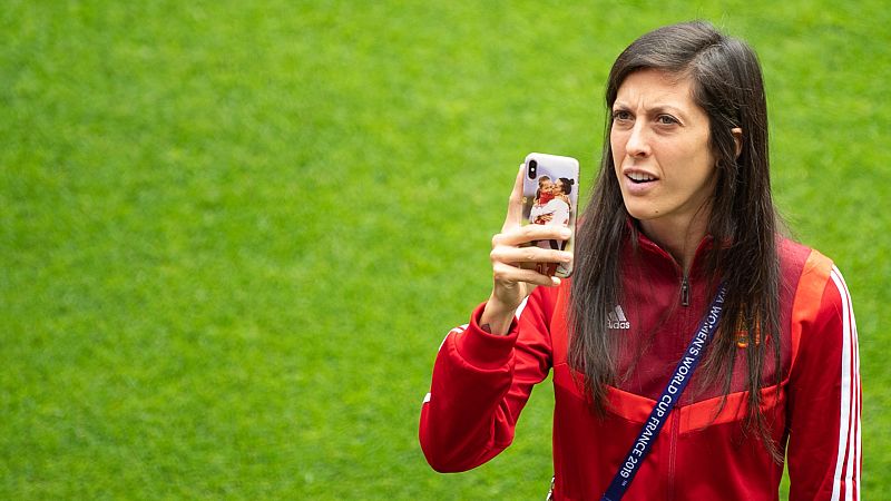 Futbolista con chaqueta roja y acreditación observa su móvil, donde se ve una imagen relacionada con un beso no consentido. Expresión seria.