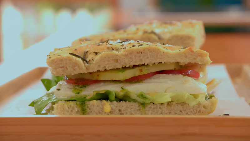 Sándwich de focaccia con tomate, mozzarella, lechuga y verduras, aderezado con hierbas y salsa verde. Pan dorado y crujiente.