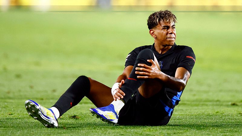 Futbolista sentado en el césped, sujetándose el pie con gesto de dolor. Lleva camiseta negra con detalles azules y botas azul y amarillo.