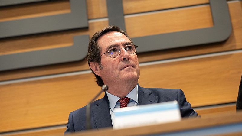 Presidente de la CEOE, en traje, sentado en una mesa, mirando hacia arriba durante una aparente rueda de prensa. Rechaza la condonación de deuda.