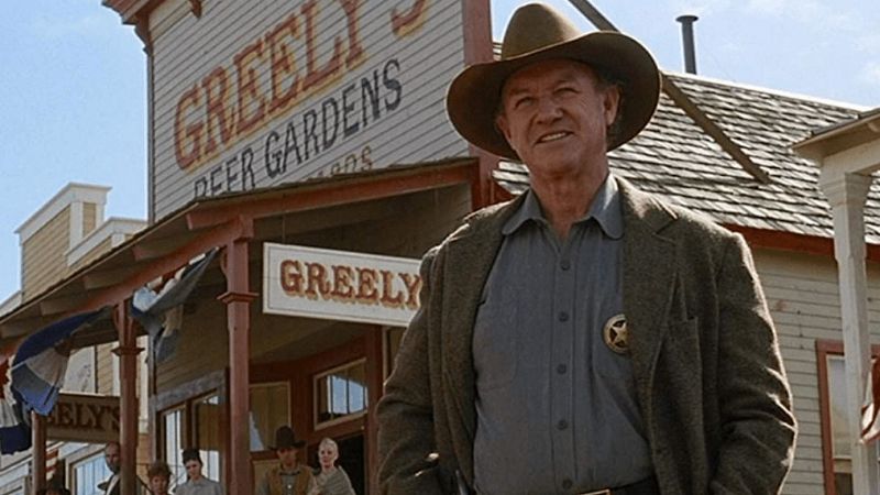 Imagen de Gene Hackman en "Sin Perdón", con atuendo de sheriff (sombrero, chaqueta marrón, camisa gris y estrella) frente a un edificio de madera con letrero "GREELY'S DEER GARDENS".