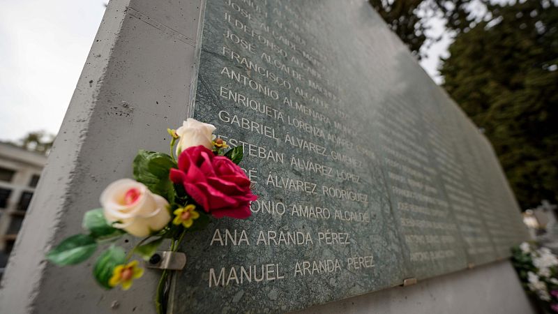 Lápida con nombres grabados en blanco sobre fondo gris verdoso, adornada con flores artificiales.  Se observan árboles y parte de otra construcción al fondo.