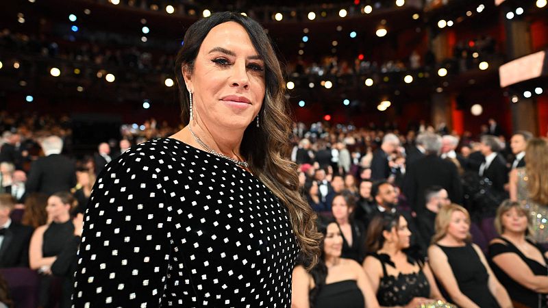 Primer plano de Karla Sofía Gascón en los Premios Oscar; viste un vestido negro con lunares blancos y sonríe ligeramente a cámara. Fondo: auditorio iluminado.