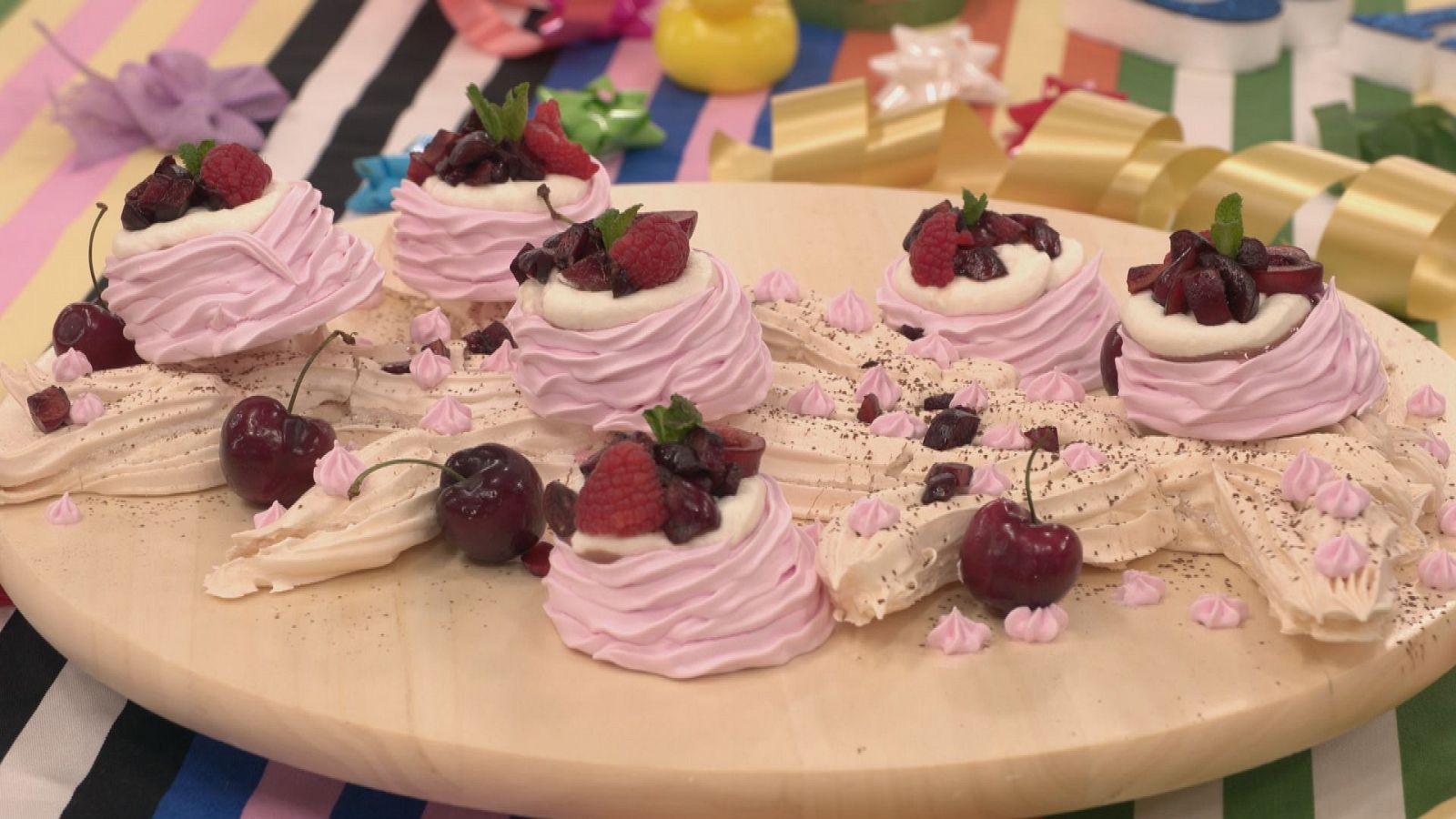 Gran pavlova con merengues rosas, frutas rojas y hojas de menta, sobre tabla de madera y mantel colorido. Base espolvoreada con cacao.
