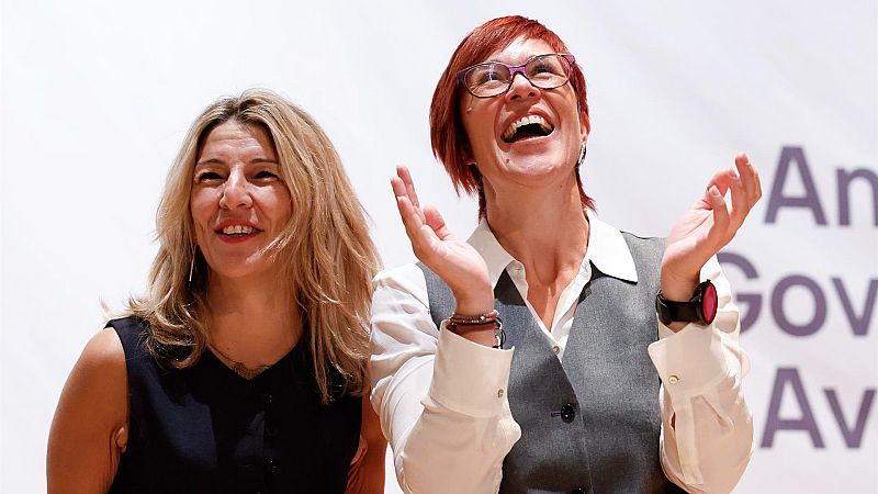 Dos mujeres sonrientes, una con blusa oscura y otra con camisa blanca y chaleco gris, celebran juntas un evento con un cartel al fondo.