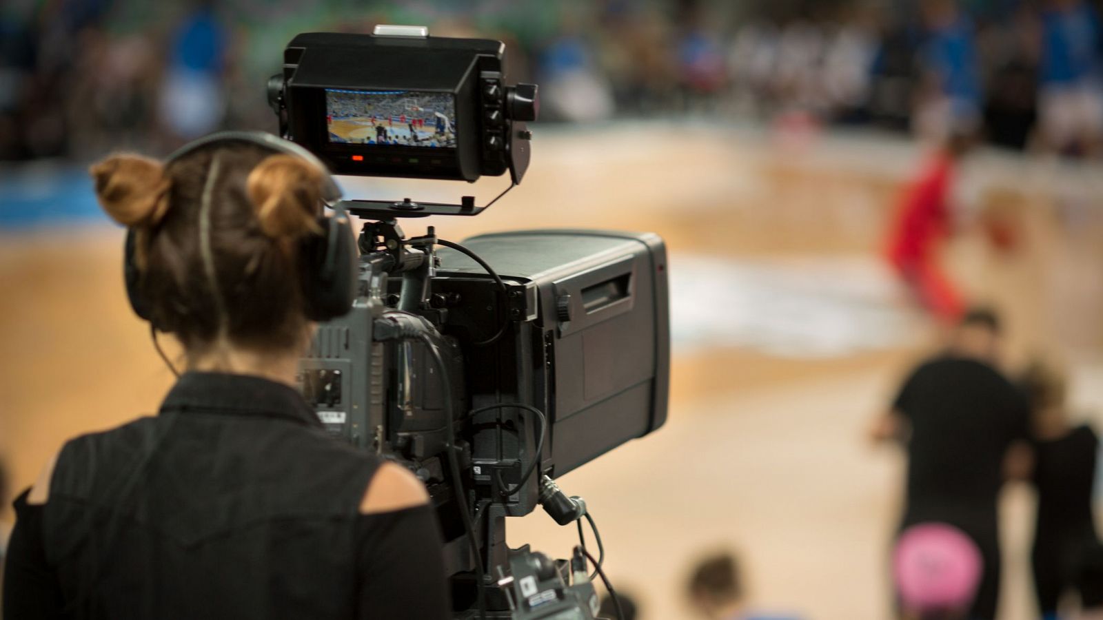 Camarógrafa grabando partido deportivo femenino; imagen enfocada en ella, jugadoras y público desenfocados; texto: "Las jóvenes recogen el testigo de las pioneras... Recordamos".