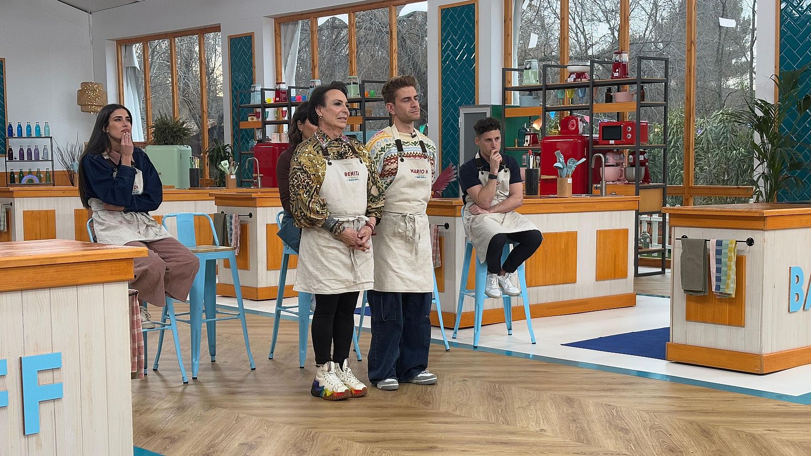 Escena de 'Bake Off: famosos al horno' con Benita y Mario Marzo (con delantales 'MARIO') ante los jueces.  Se aprecian otros concursantes y detalles del set de grabación.