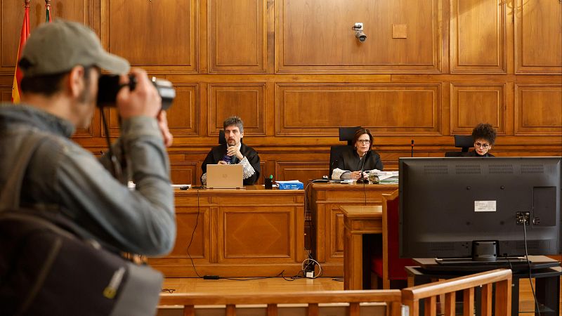 Juicio en una sala de madera. Tres personas (un hombre con toga y dos mujeres) sentadas en una mesa con un portátil y documentos. Un fotógrafo toma imágenes.